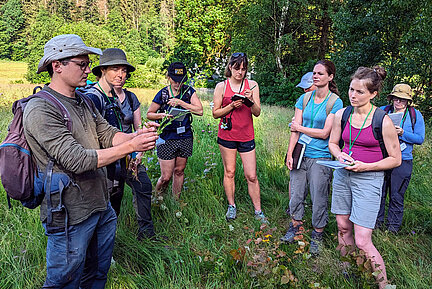 Teilnehmerinnen und Teilnehmer einer Botanik-Exkursion. (Foto: Anja Gellert) Eine Gruppe Menschen steht draußen im Kreis und beobachtet Gräser. und